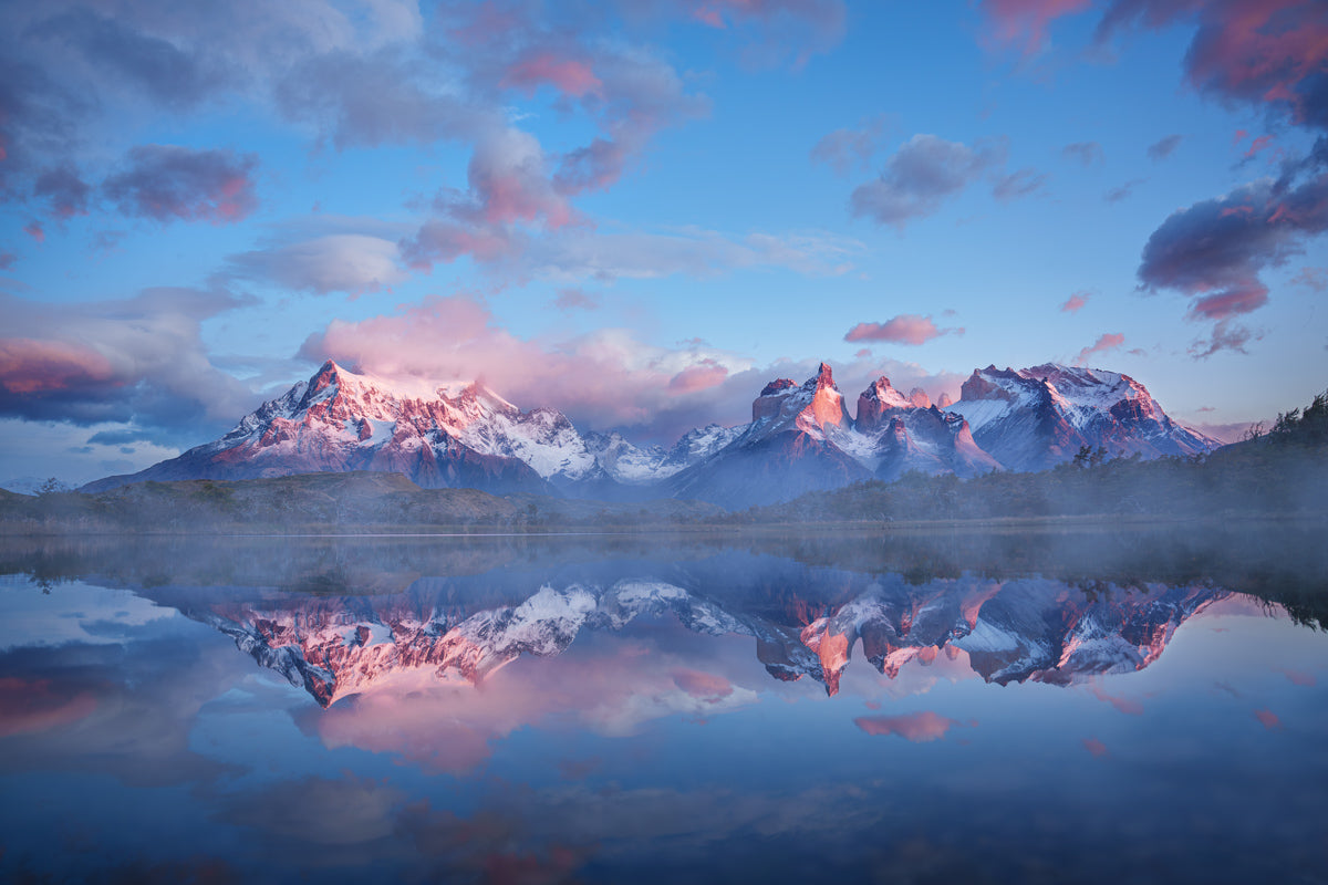 A scenic landscape photograph of snow-capped mountains reflected in a calm lake under a blue sky with pink clouds.